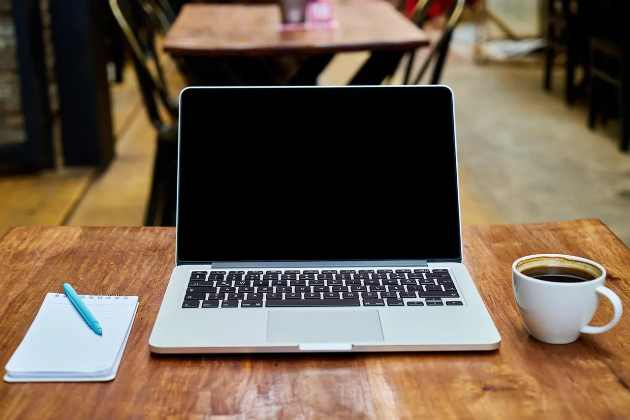 Laptop con schermo spento su un tavolo di legno, accanto a una tazza di caffè e a un taccuino con penna azzurra, in un caffè.
