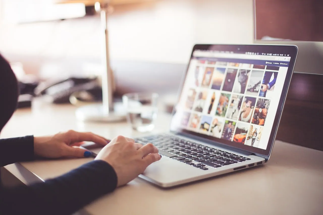 Due mani su un laptop aperto che mostra una griglia di miniature di foto, su una scrivania con un bicchiere d'acqua sullo sfondo.