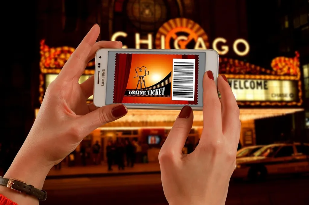 Due mani con smalto rosso tengono uno smartphone che mostra un biglietto online con codice a barre, davanti all'insegna luminosa del teatro Chicago di sera.
