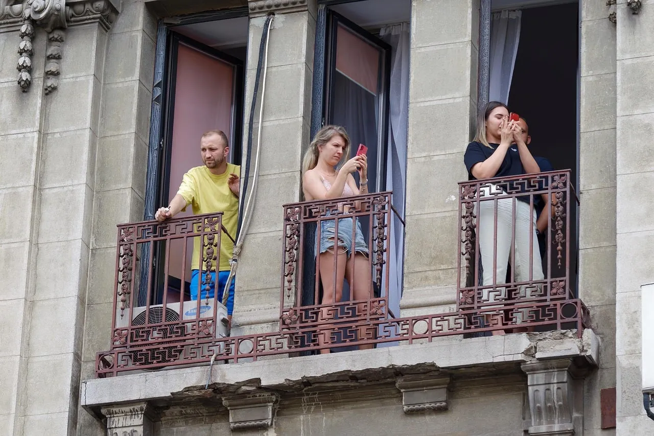 Tre persone su un balcone in ferro battuto di un palazzo antico guardano in basso; due di loro tengono in mano uno smartphone.