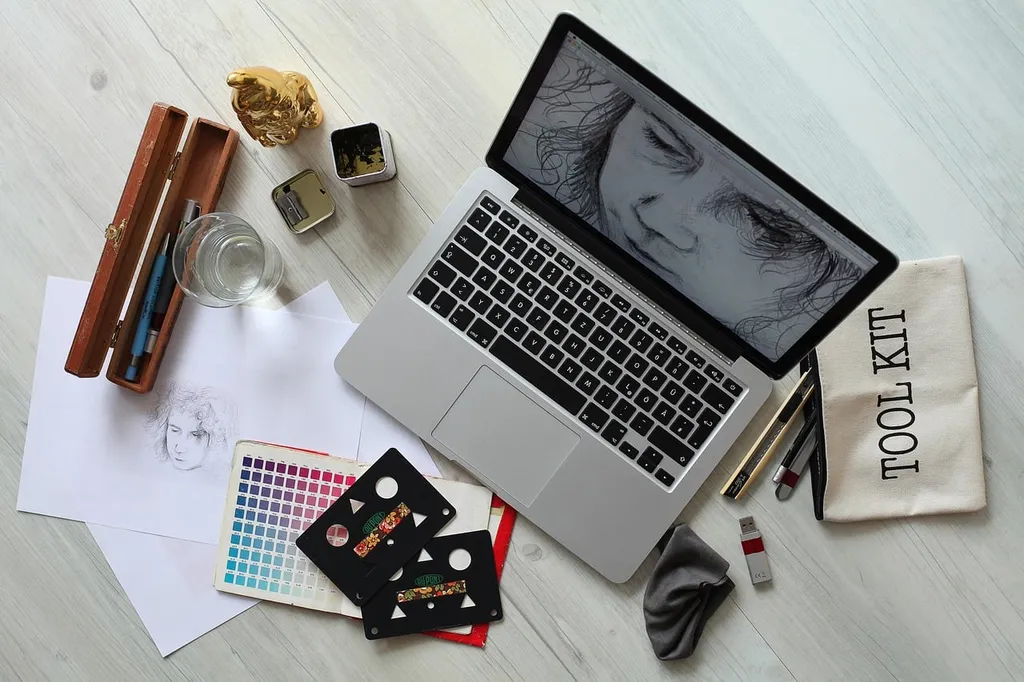 Scrivania vista dall'alto con laptop che mostra un disegno di volto, campionario colori, schizzi su carta, bicchiere e una trousse "Tool Kit".