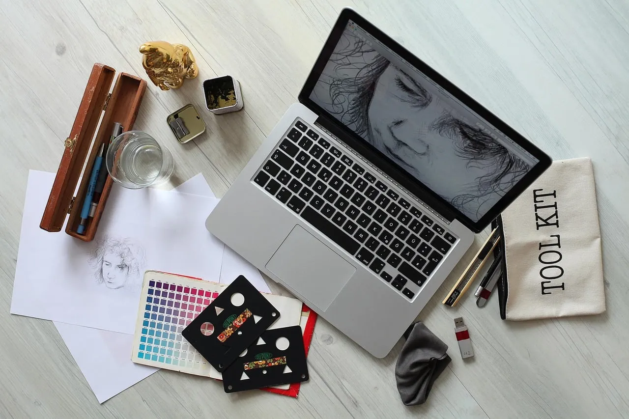 Scrivania vista dall'alto con laptop che mostra un disegno di volto, campionario colori, schizzi su carta, bicchiere e una trousse "Tool Kit".